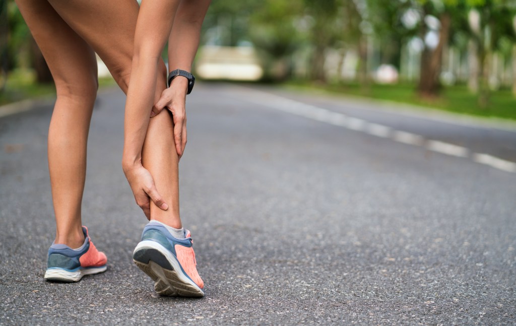 woman holds her calf leg as if injured, while standing on a pavement