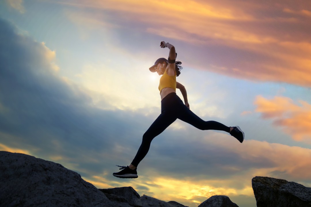 Female runner jumping over a mountain ridge at sunset