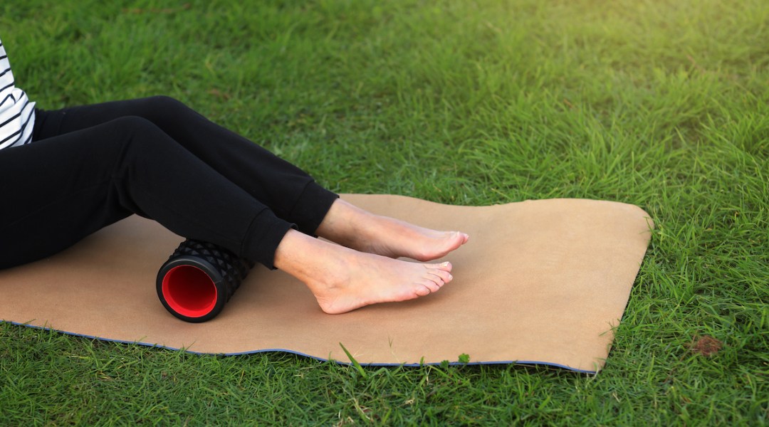 Unrecognizable woman warms up and works out the fascia of the calf muscles with a sport massage foam roller outdoors on green grass