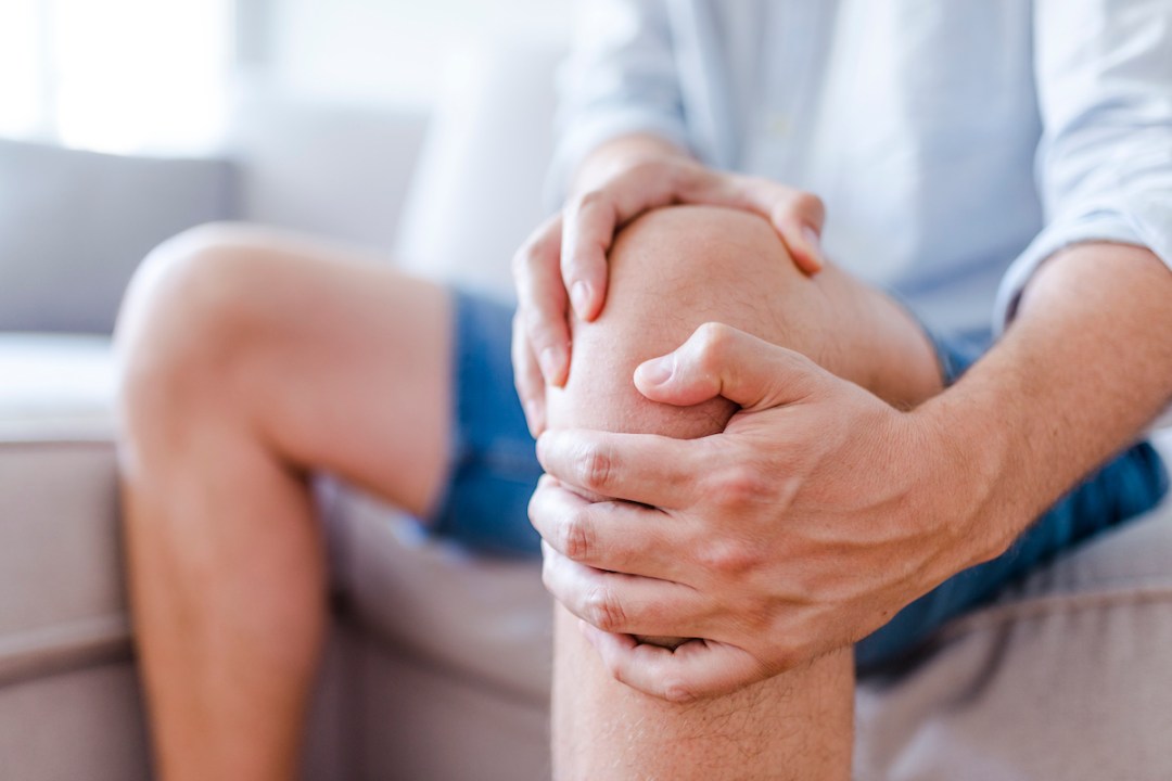 Man suffering from knee pain sitting on sofa, massaging his painful knee