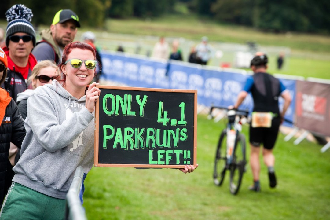 Smiling woman wearing sunglasses holding up a motivational sign during a triathlon race day. The caption reads 'Only 4.1 park runs left!!'