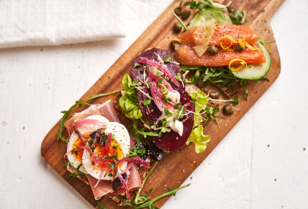 Overhead view of three open sandwiches, aka Smorrebrods, arranged on a brown chopping board