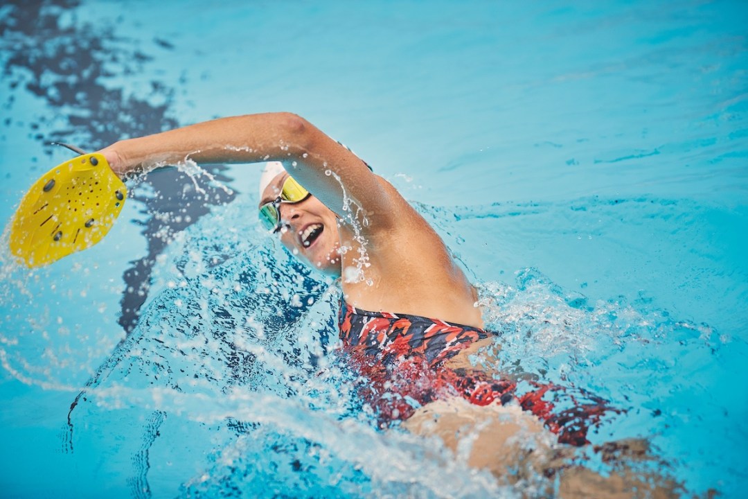 Female swimmer training in pool, breathing to one side, wearing yellow paddle on left hand as it lifts out of the water