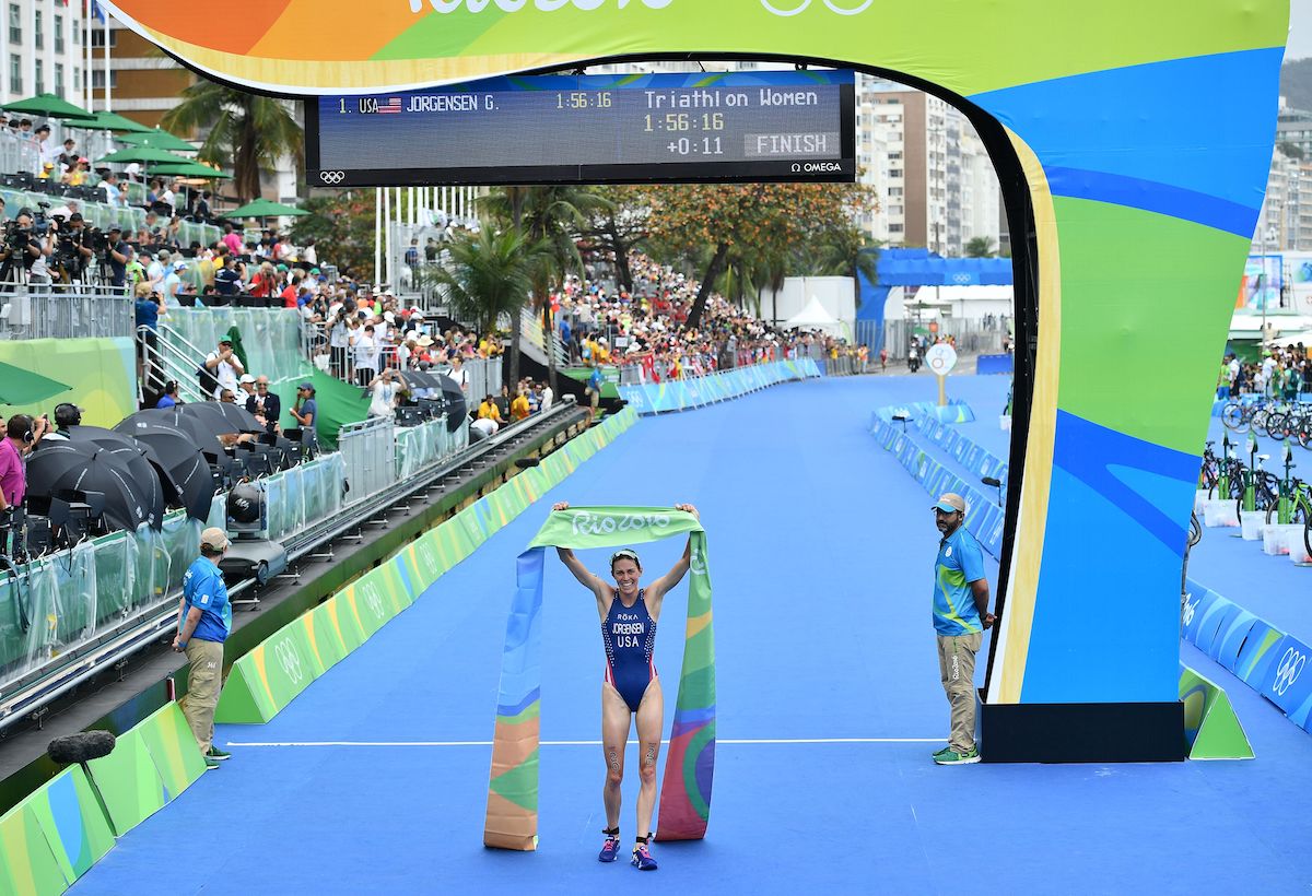 USA's Gwen Jorgensen crosses the line at the 2016 Rio Olympic Game to win gold, holding the finisher's tape aloft, with crowds and media on the left hand of the image