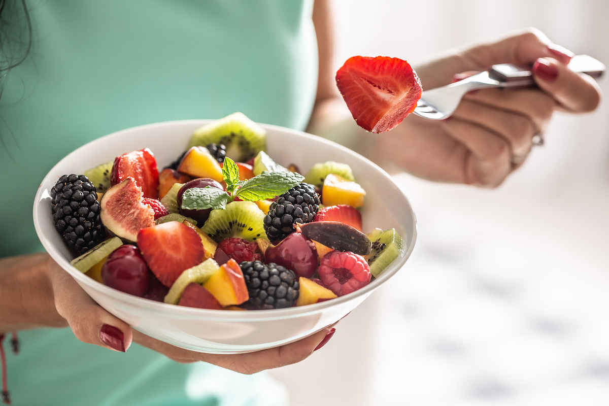 A close-up of a woman's hands holding a bowl full of fruit with a fork in her left hand holding a strawberry