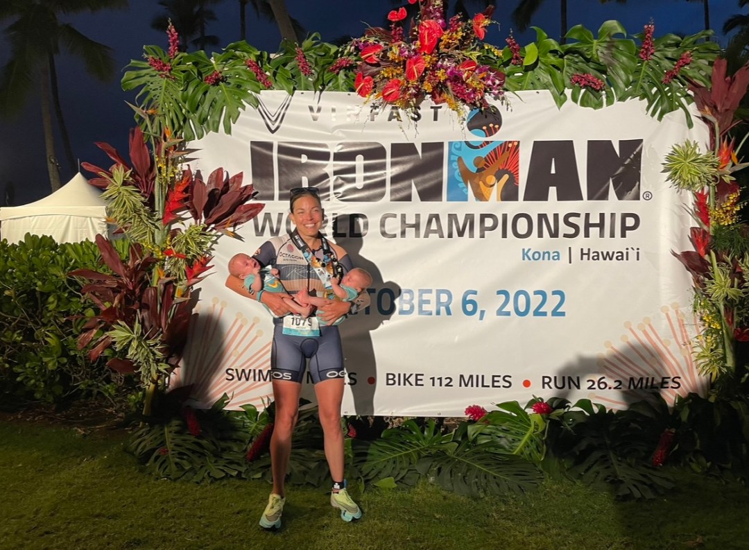 Jacqui Giuliano poses in front of a large banner at the 2022 Ironman World Champs, holding her twin boys in each arm