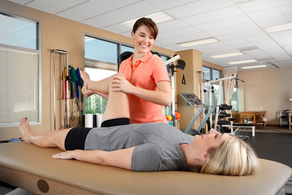 A smiling female physical therapist evaluating the range of motion of a young female patient's hip as the patient lies on a treatment table.  Scene is inside a typical physical therapy clinic with several pieces of exercise equipment in the background. Physical therapist is in her early 30's and the patient is in her early 20's.