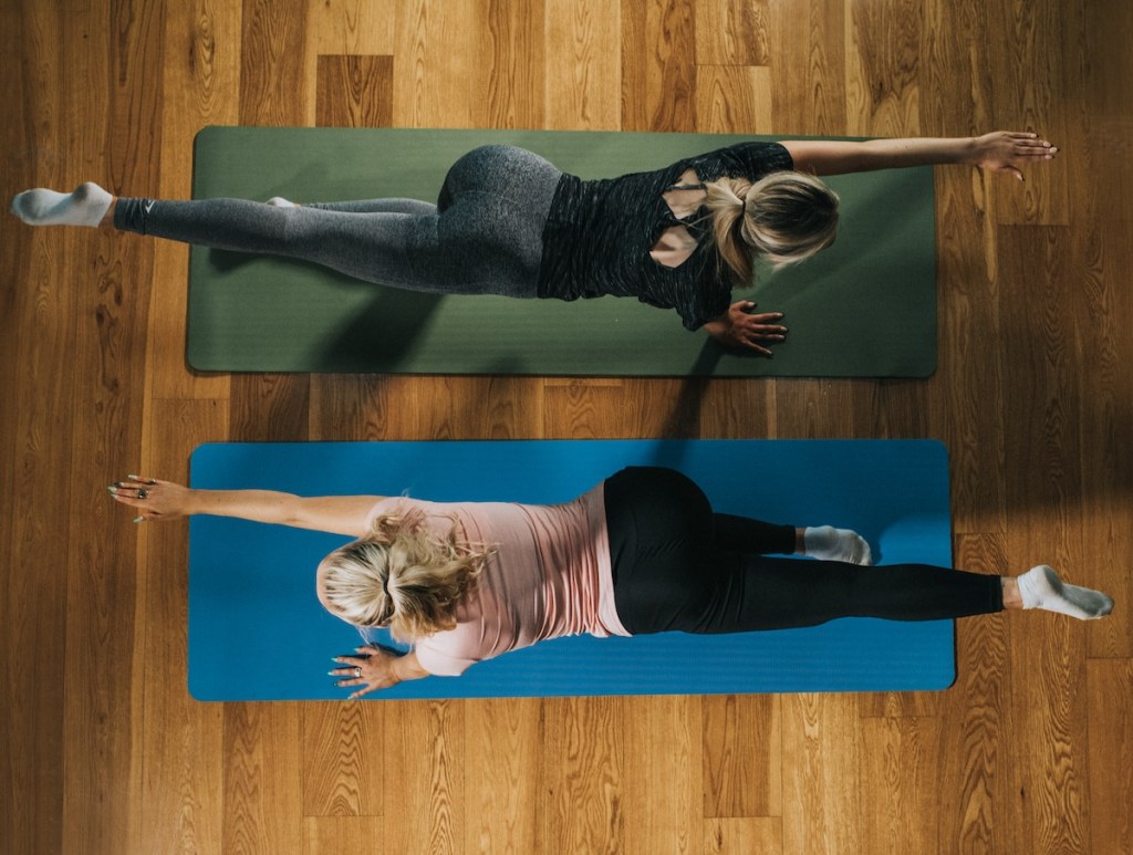 Two young woman in their twenties perform yoga on yoga mats on a wooden floor. They perform the 'superman' pose with one leg and one arm outstretched.