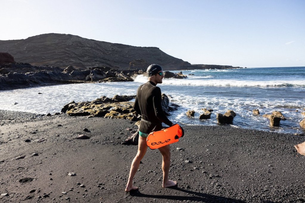 Male swimmer walking to the sea in a neoprene t-shirt carrying a tow float